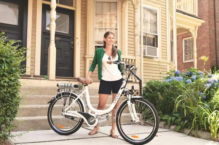 Why Renting an E-Bike is the Best Way to See Vancouver Woman with an electric bicycle in front of a suburban home, suggesting eco-friendly lifestyle and urban commuting.
